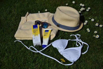 Einige Badesachen liegen auf der Wiese: ein Handtuch, die Sonnen-Hautmilch von Linola, ein Sonnenhut und ein Bikini.